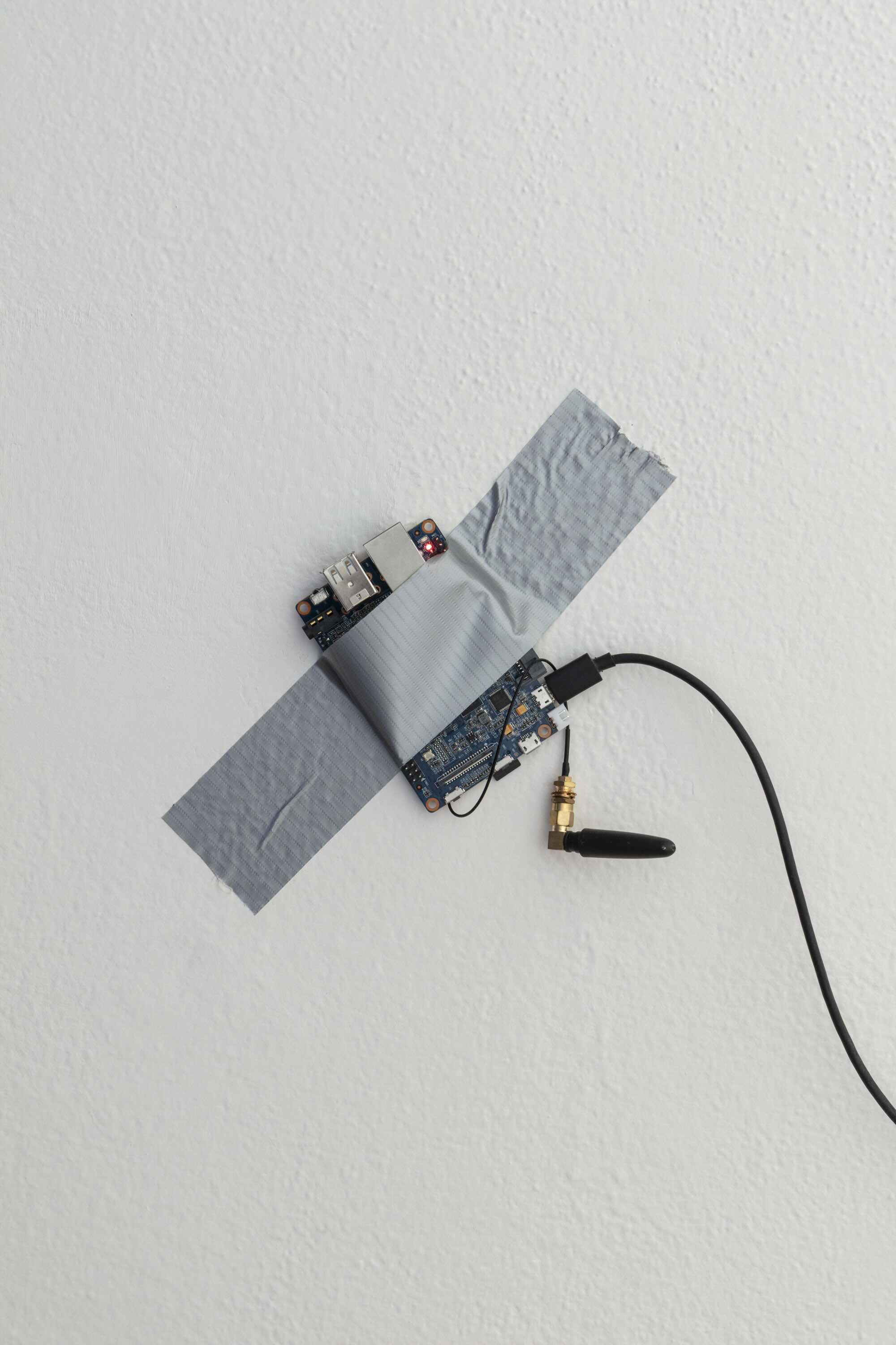 Single board computer BananaPi M1+ duct-taped to a white wall, with a black USB powercable, lighted red LED diode and a mini wifi antenna hanging.